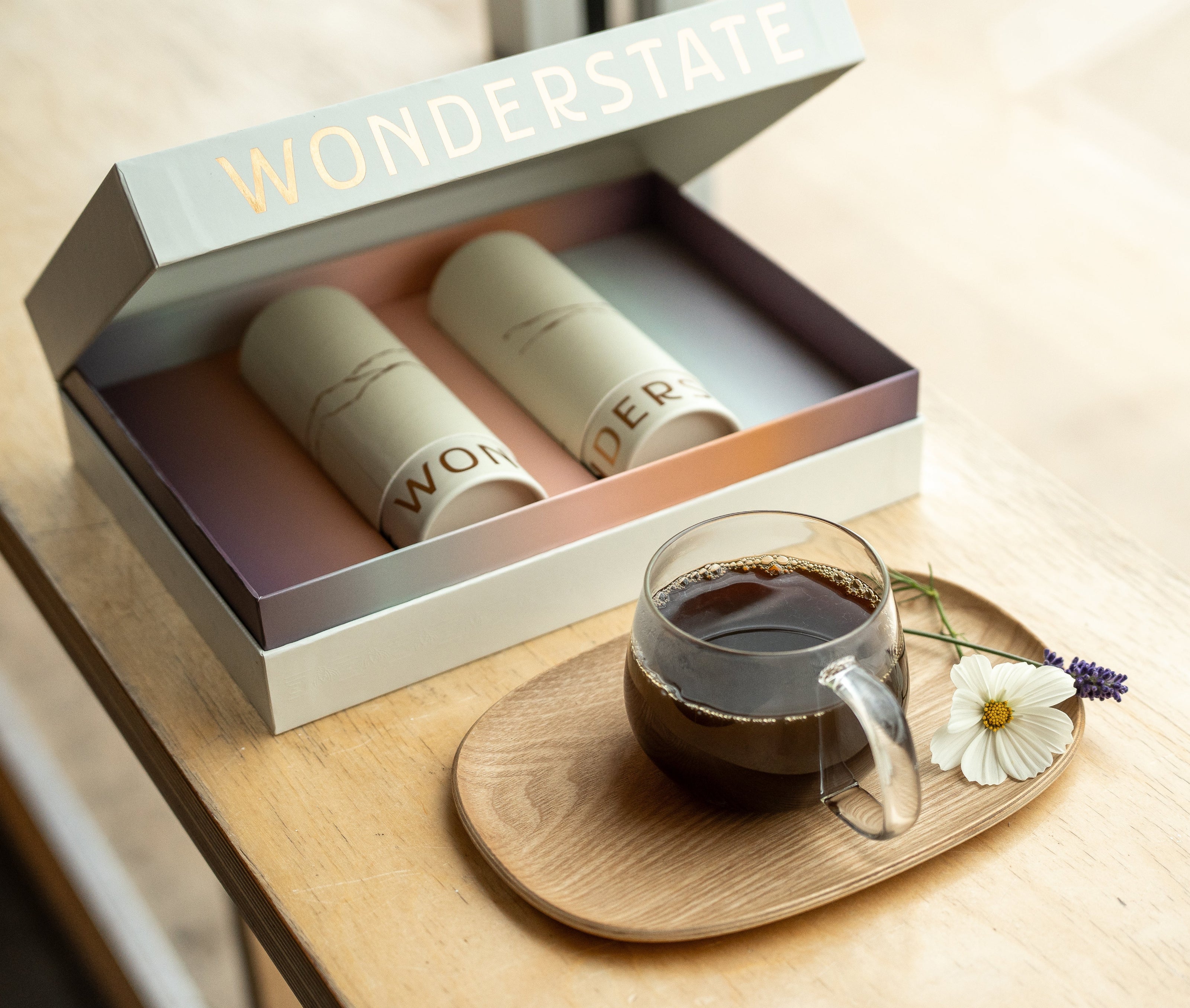 Box with two coffee tubes inside next to a cup of coffee on a tray with flowers.